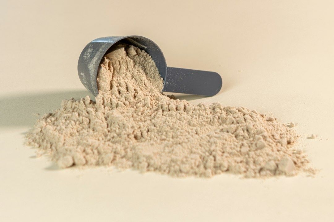 Black scoop spilling pale protein powder onto a beige surface